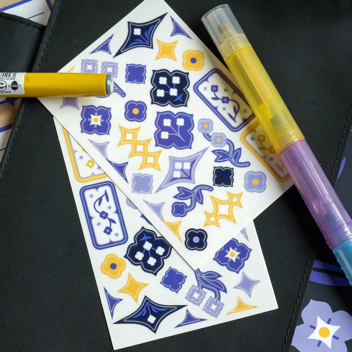 A set of decorative stickers featuring speedwell flower illustrations in blue, purple, and yellow are displayed against the cover of a black notebook. Yellow, blue, and purple pens and highlighters surround the scene.