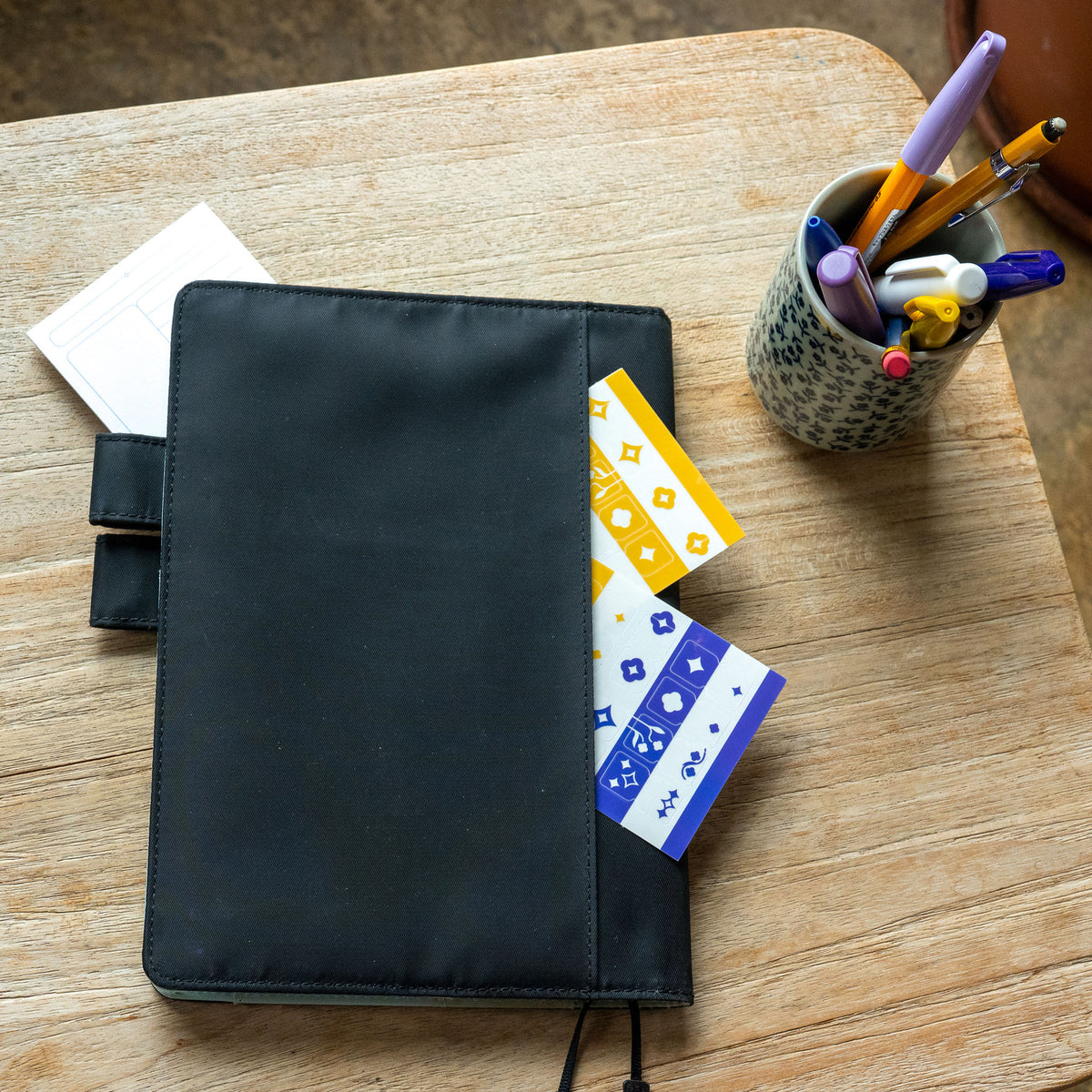 A planner with a black fabric cover lays on a wooden side table. The Archivist Sticky Note and Tab Divider Stickers are tucked into the the planner's back pocket. A pencil cup full of purple, yellow, and blue pencils sits to the right.