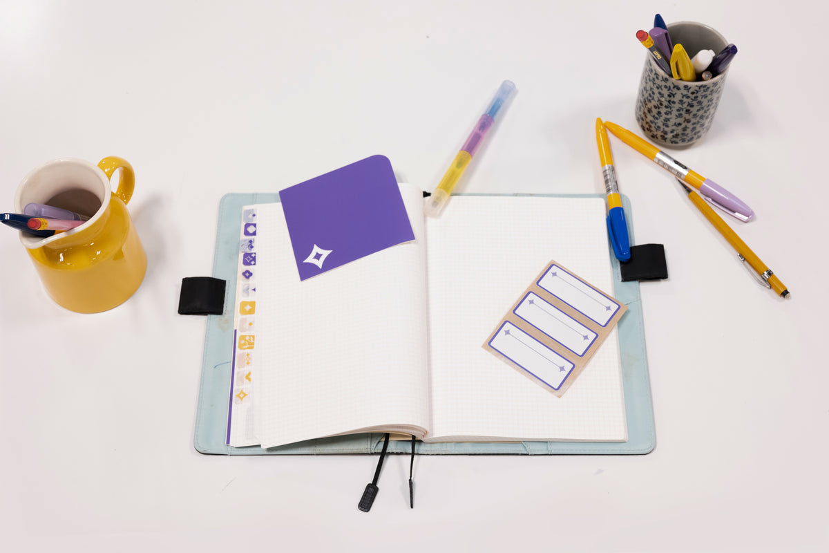 An open notebook with Tab Divider Stickers, Pocket Sticker and Nameplate Stickers surrounded by multiple pencil cups with pens, pencils, and markers in yellow, blue, and purple.