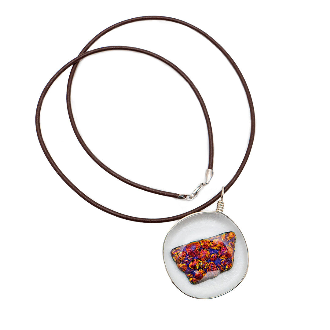 ember glass pendant - a brightly colored & irregularly shaped piece inside a round glass pendant - is attached to a coiled brown leather cord against a white background.