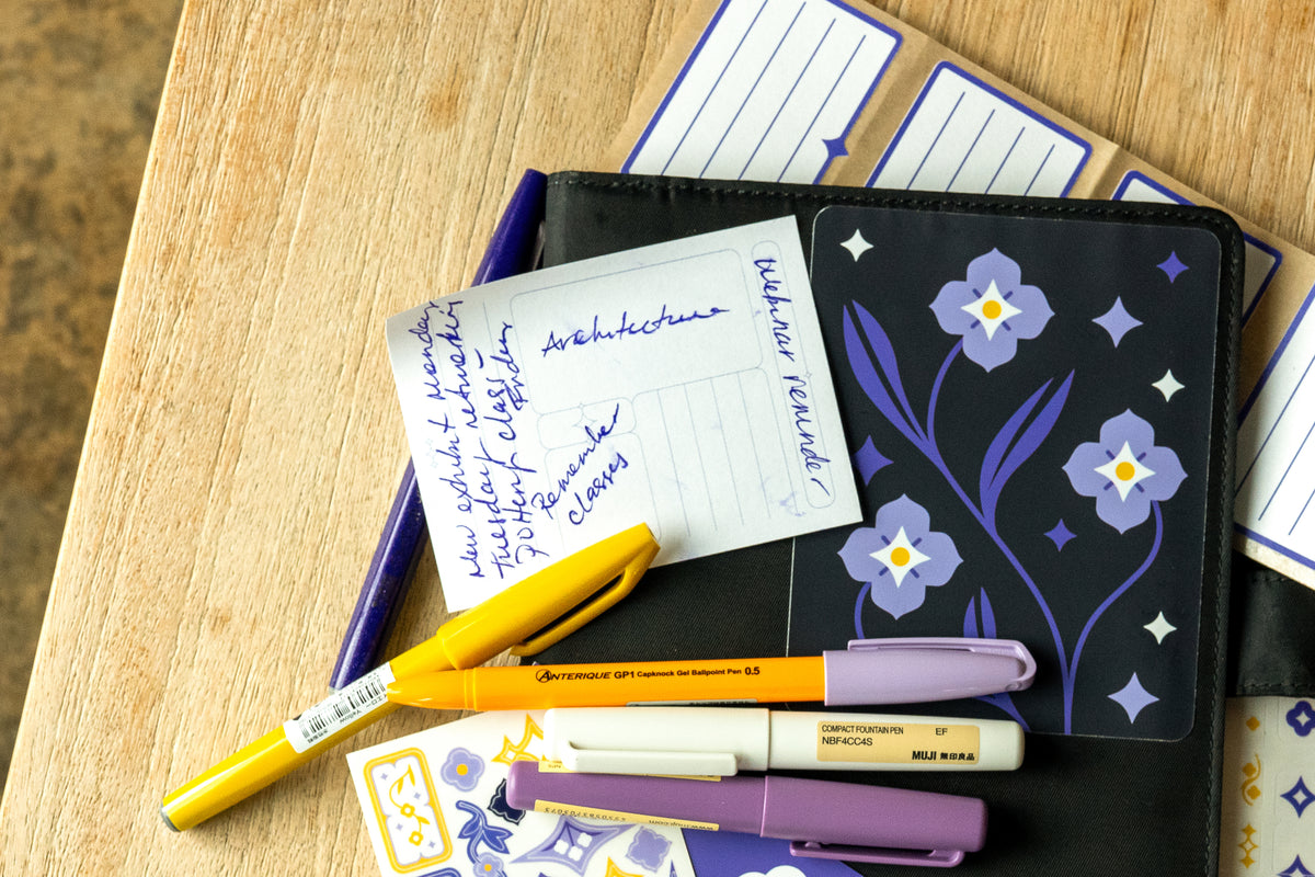 A close-up flat lay of the Speedwell Stationery on a wooden table, featuring the Cover Sticker, Archivist Sticky Note, Sticker Sheets, and Nameplate Stickers, surrounded by yellow and purple pens.