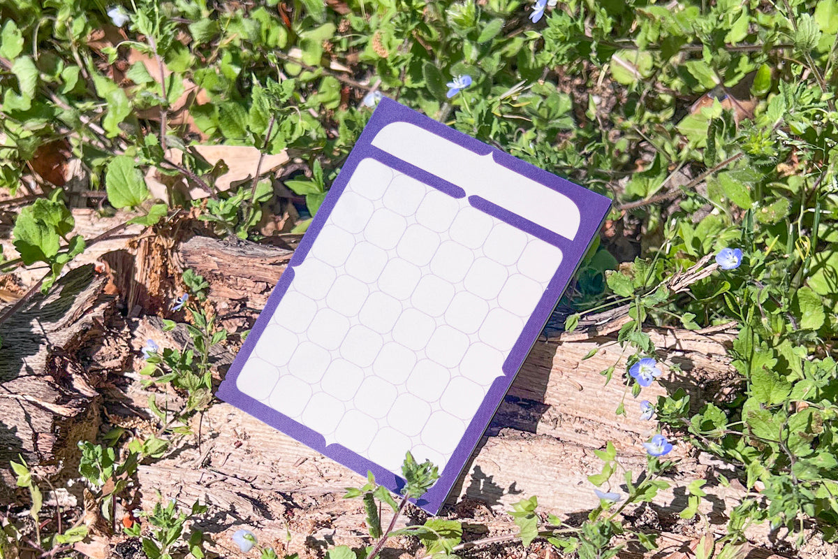 The Calendar Sticky Note pad photographed against the vibrant green and indigo of living Speedwell flowers.