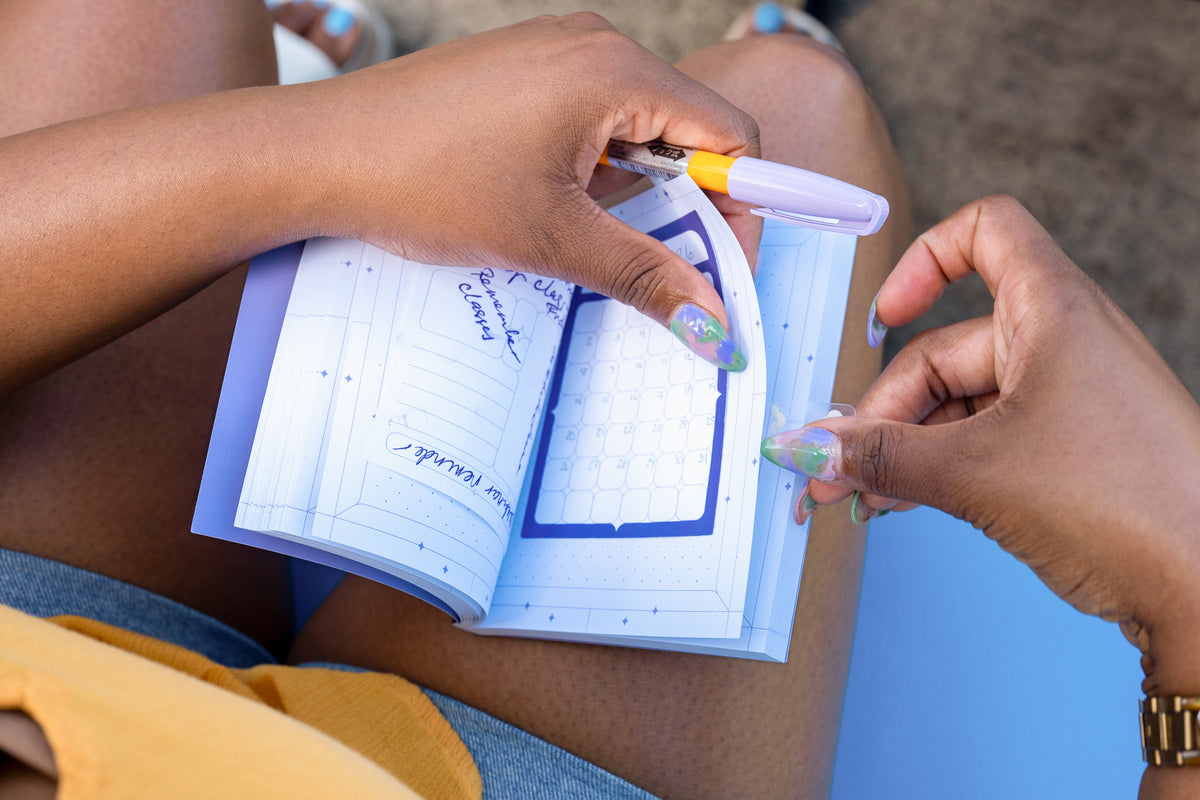 A close-up lifestyle shot of a person's hands holding an open pocket notebook - on the left page is the Archivist Sticky Note and on the right page is the Calendar Sticky Note. The model has filled out the sticky notes with a yellow and lavender pen.