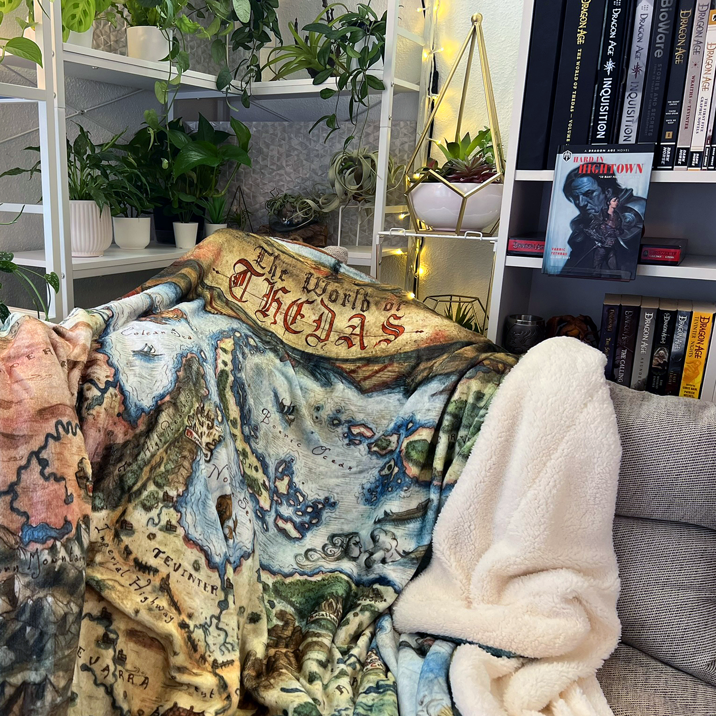 The Thedas Map Blanket draped over a sofa with a complete collection of Dragon Age books in the background.