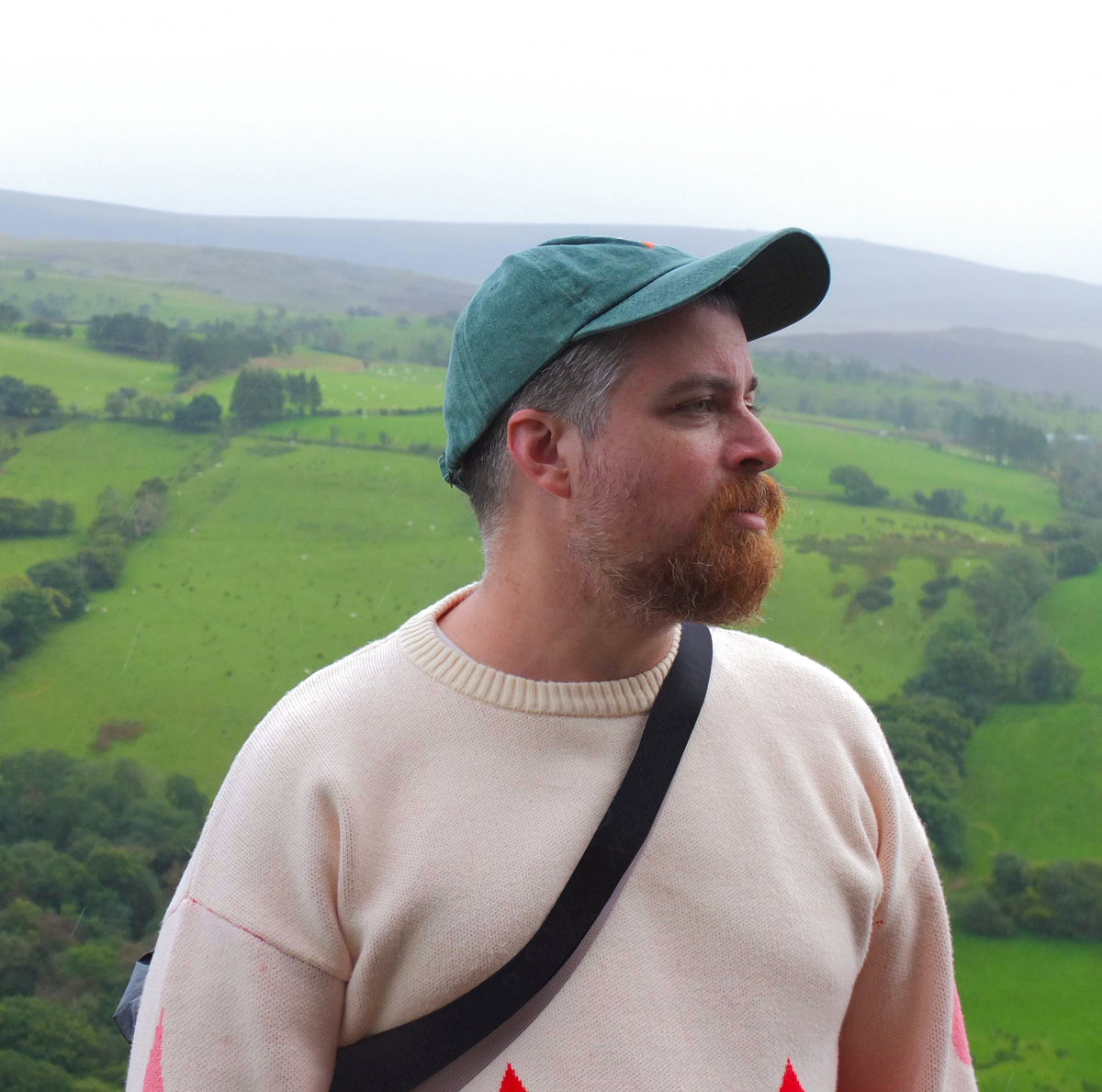 A bearded man wearing a green cap and a cream-colored sweater looks out over the rolling green hills of Wales.