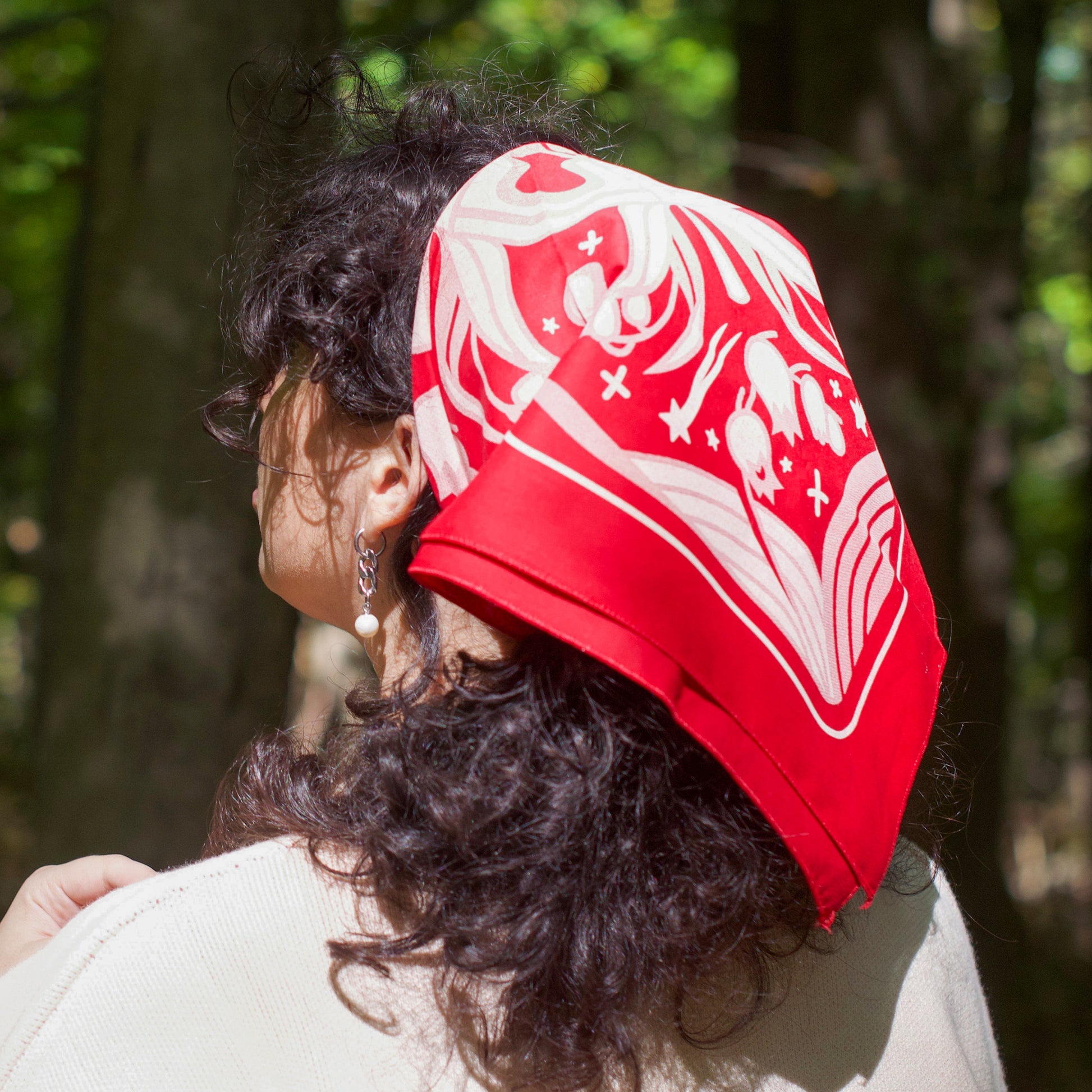 A model, facing away from the viewer, is wearing the bandana as a headscarf. A corner is pointed downward across the back of her hair, which shows the print of the lily of the valley plant facing upright.