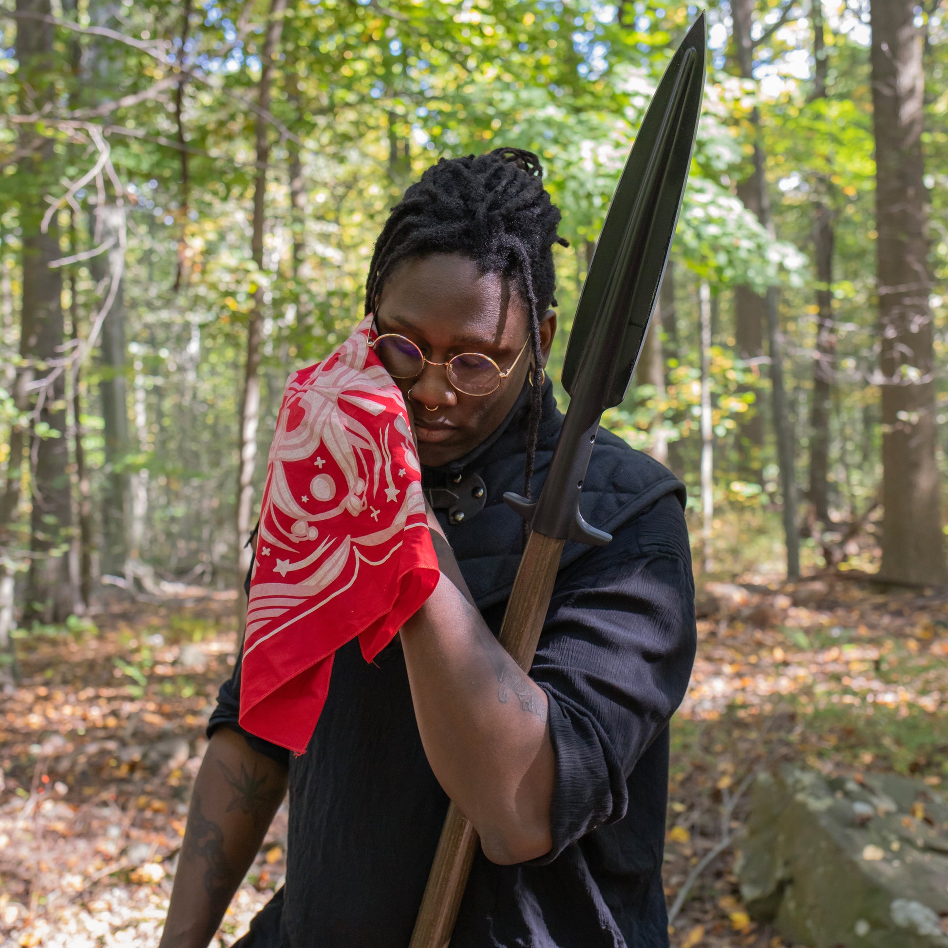 A model is leaning on their spear in the forest, eyes closed, and wiping their brow with the Sacred Blood Bandana.