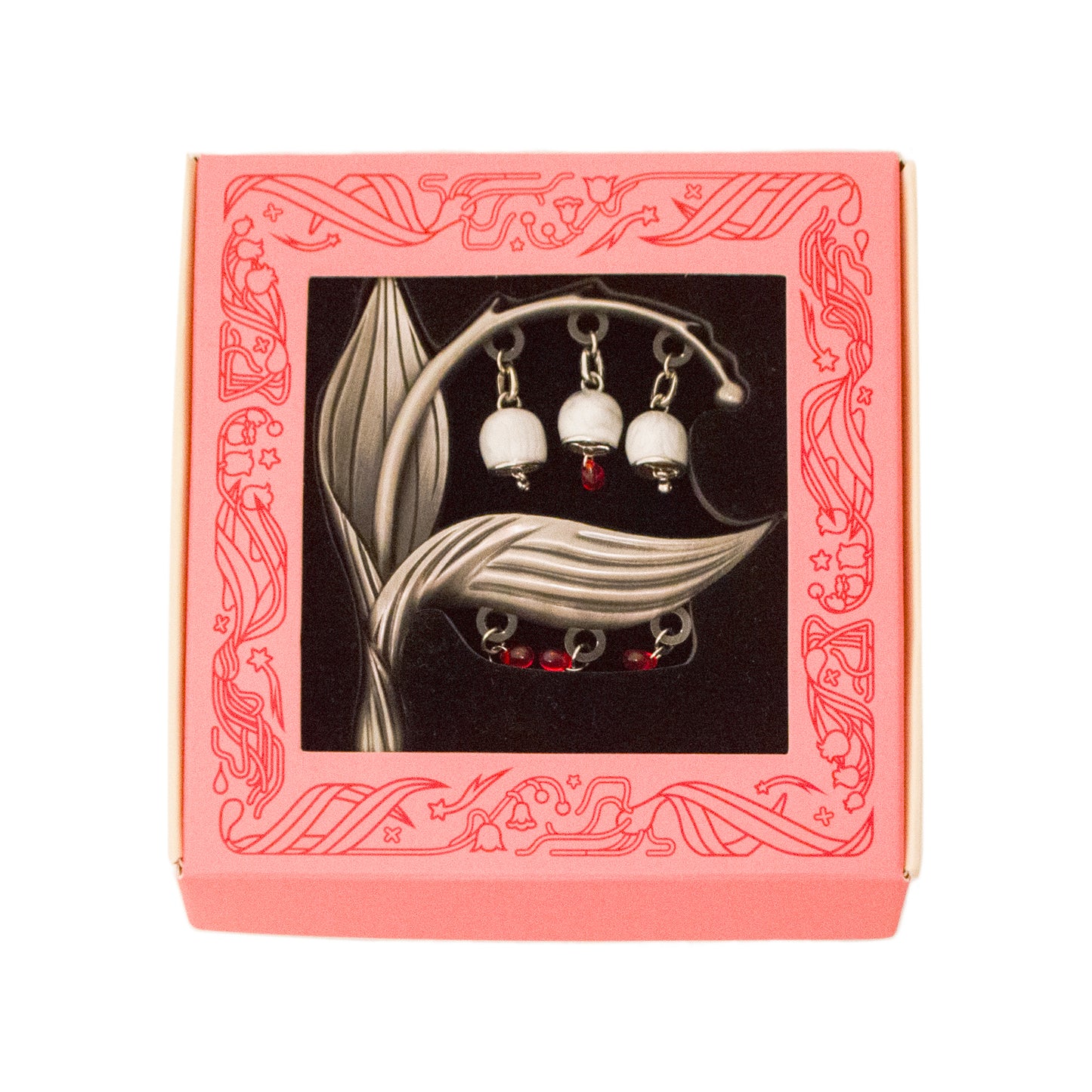 The Sacred Blood Pin is packed in a light coral pink window box with black foam. Illustrations of lily of the valley and stars adorn the border frame of the box around the window.