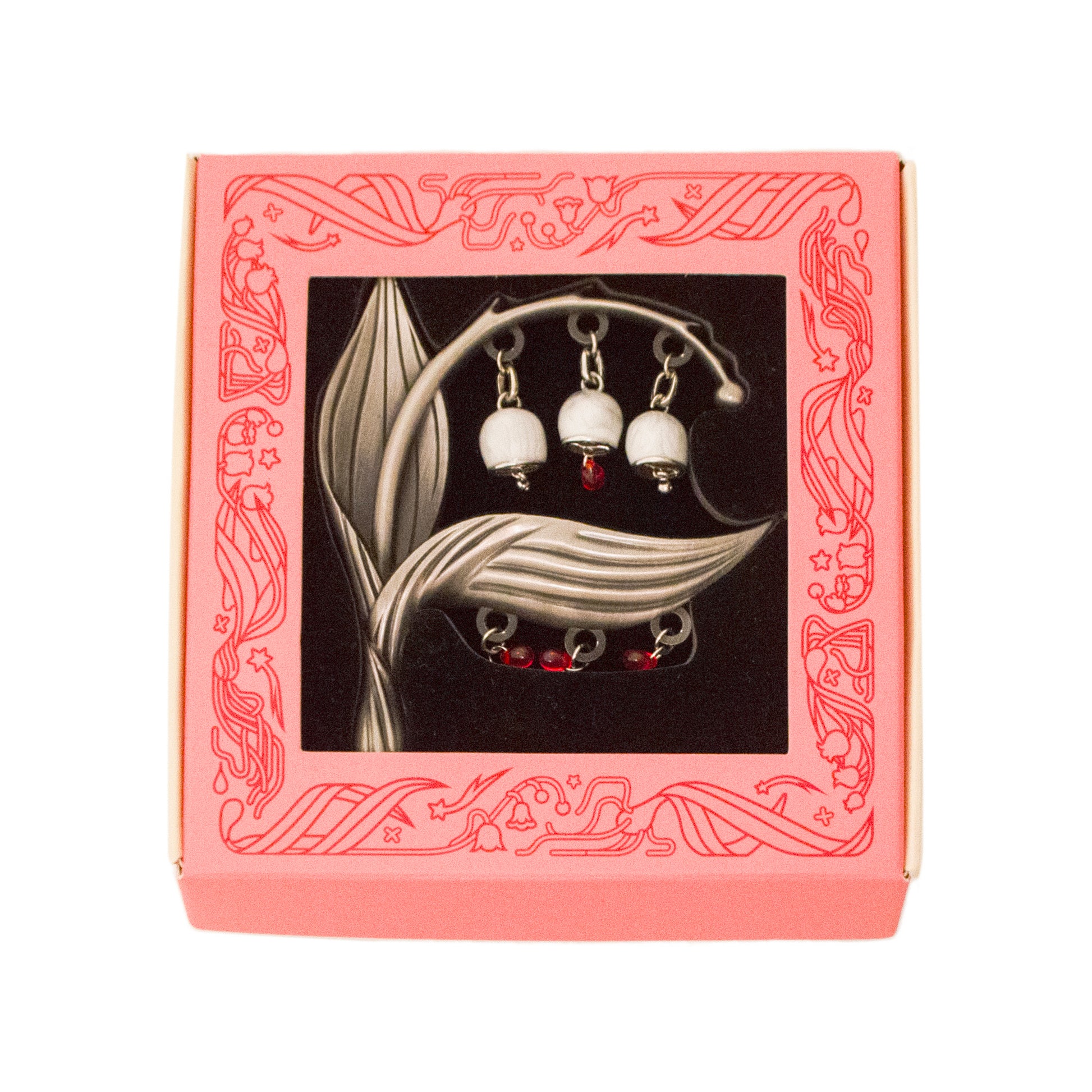 The Sacred Blood Pin is packed in a light coral pink window box with black foam. Illustrations of lily of the valley and stars adorn the border frame of the box around the window.