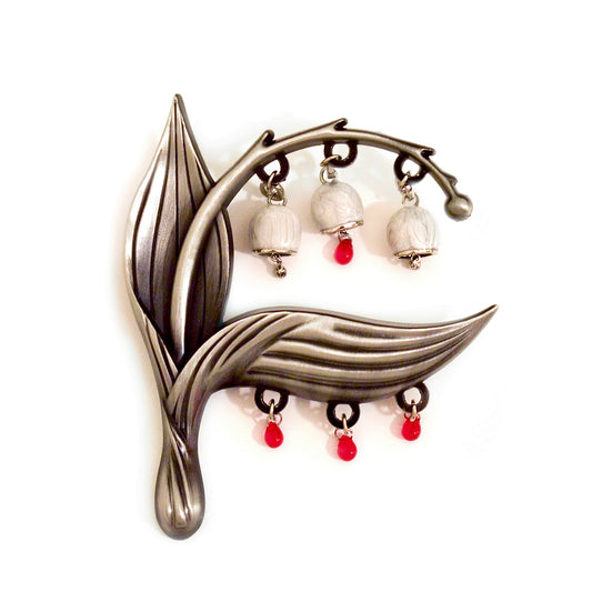 A product photo of the Sacred Blood pin, a curved lily of the valley plant in the color of warm silver. Hanging from the plant stem are three pearlized bell-shaped flowers. The middle lily has a red teardrop shaped bead hanging from it, as does the bottom, horizontally oriented leaf of the plant.
