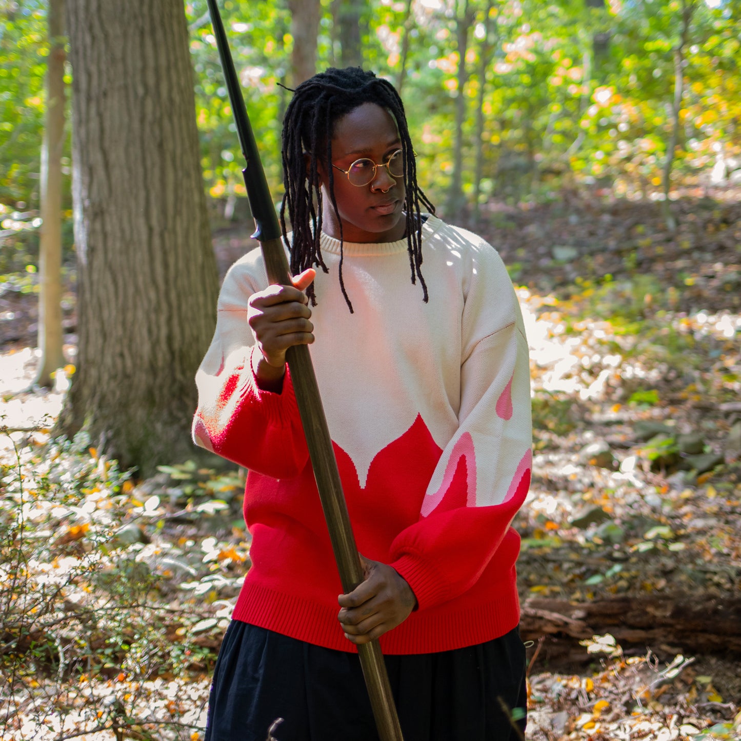 A model wearing the Sacred Blood sweater, holding a spear in a forest setting. The sweater extends to the model's hip and boldly stands out against the greenery.