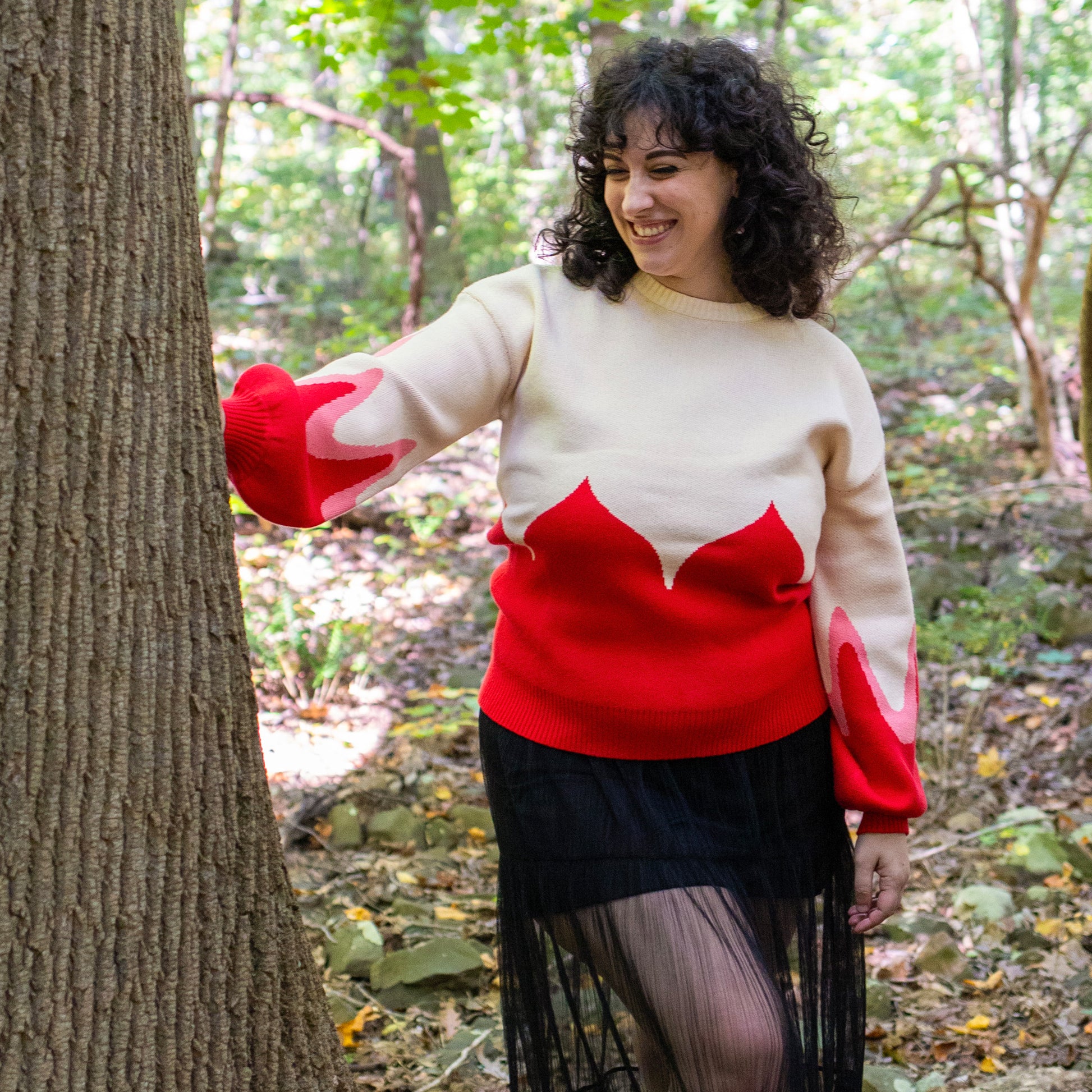 Another model wearing the Sacred Blood sweater, peeking out from behind a tree in the forest. 