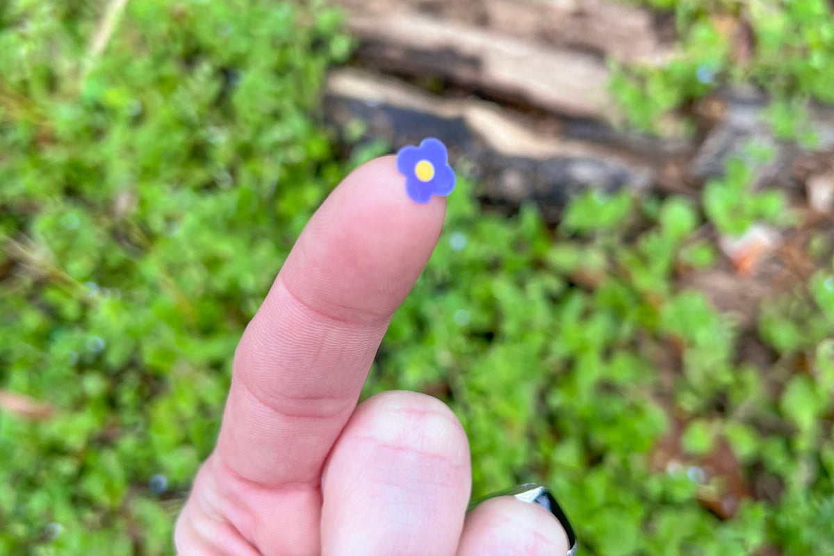 A single Speedwell Art Sticker depicting the iconic speedwell flower is photographed on the tip of a person's finger against vibrant green foliage.