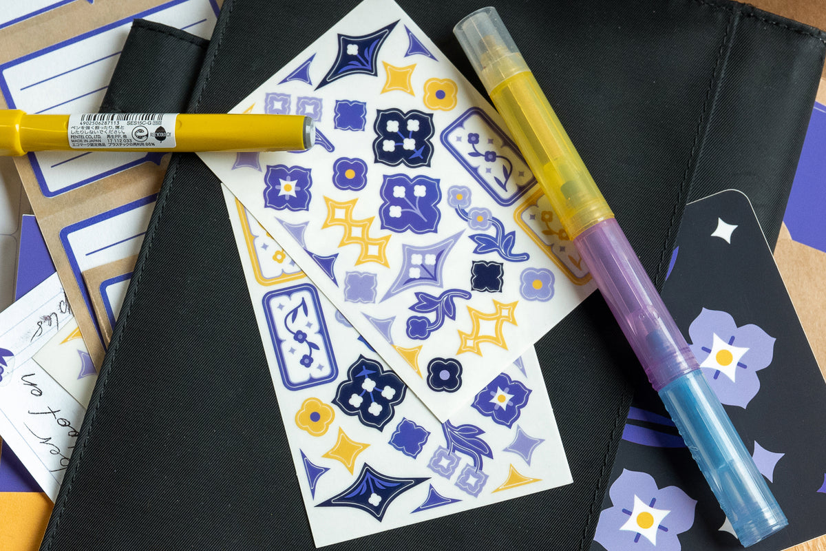 A set of decorative stickers featuring speedwell flower illustrations in blue, purple, and yellow are displayed against the cover of a black notebook. Yellow, blue, and purple pens and highlighters surround the scene.