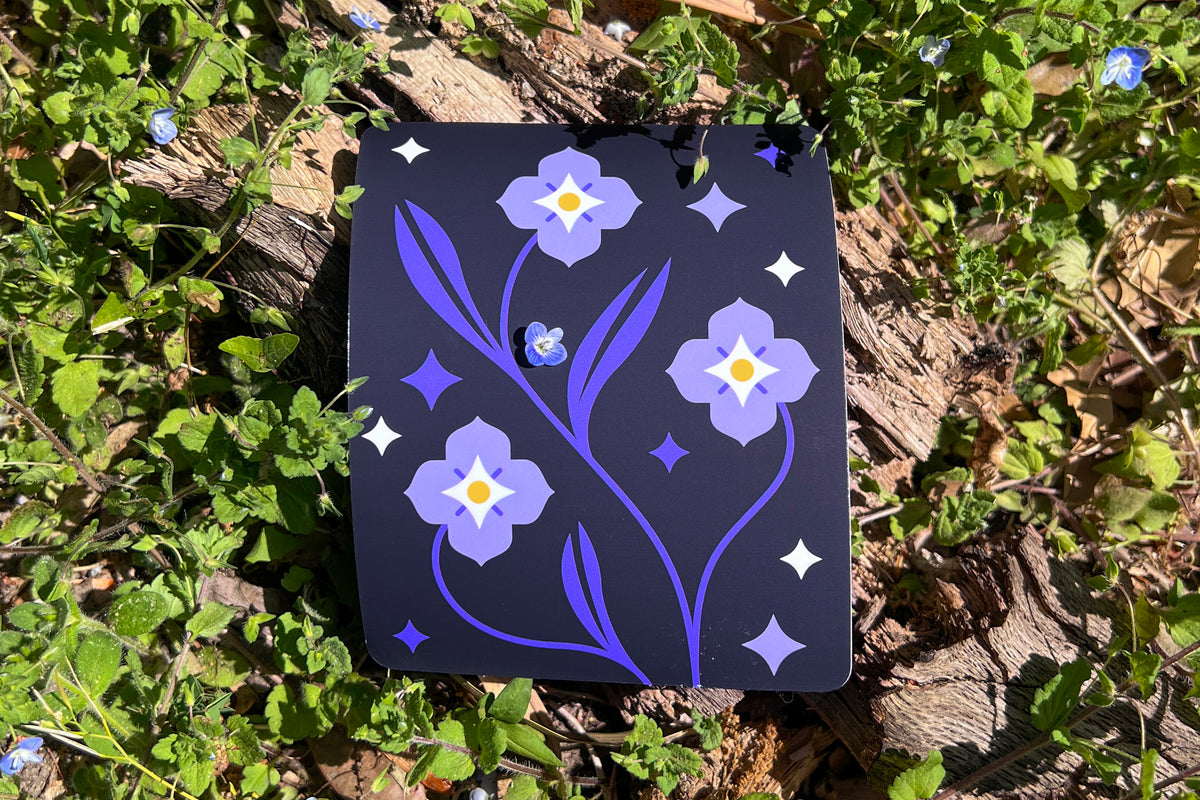 A vinyl sticker depicts Speedwell flowers in a modern and clean illustrative style with indigo, white, and yellow colors. The sticker is photographed amongst a background of the same living flowers. A single miniature speedwell blossom rests upon the sticker.