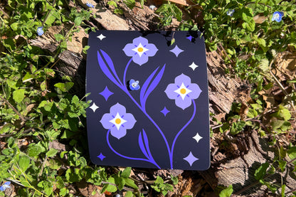 A vinyl sticker depicts Speedwell flowers in a modern and clean illustrative style with indigo, white, and yellow colors. The sticker is photographed amongst a background of the same living flowers. A single miniature speedwell blossom rests upon the sticker.