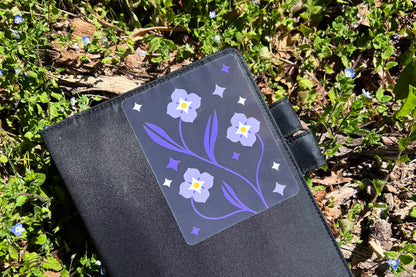 A black fabric notebook pictured amongst Speedwell flowers with a vinyl sticker on the cover depicting the same species of flowers in a modern and clean illustrative style with indigo, white, and yellow colors.