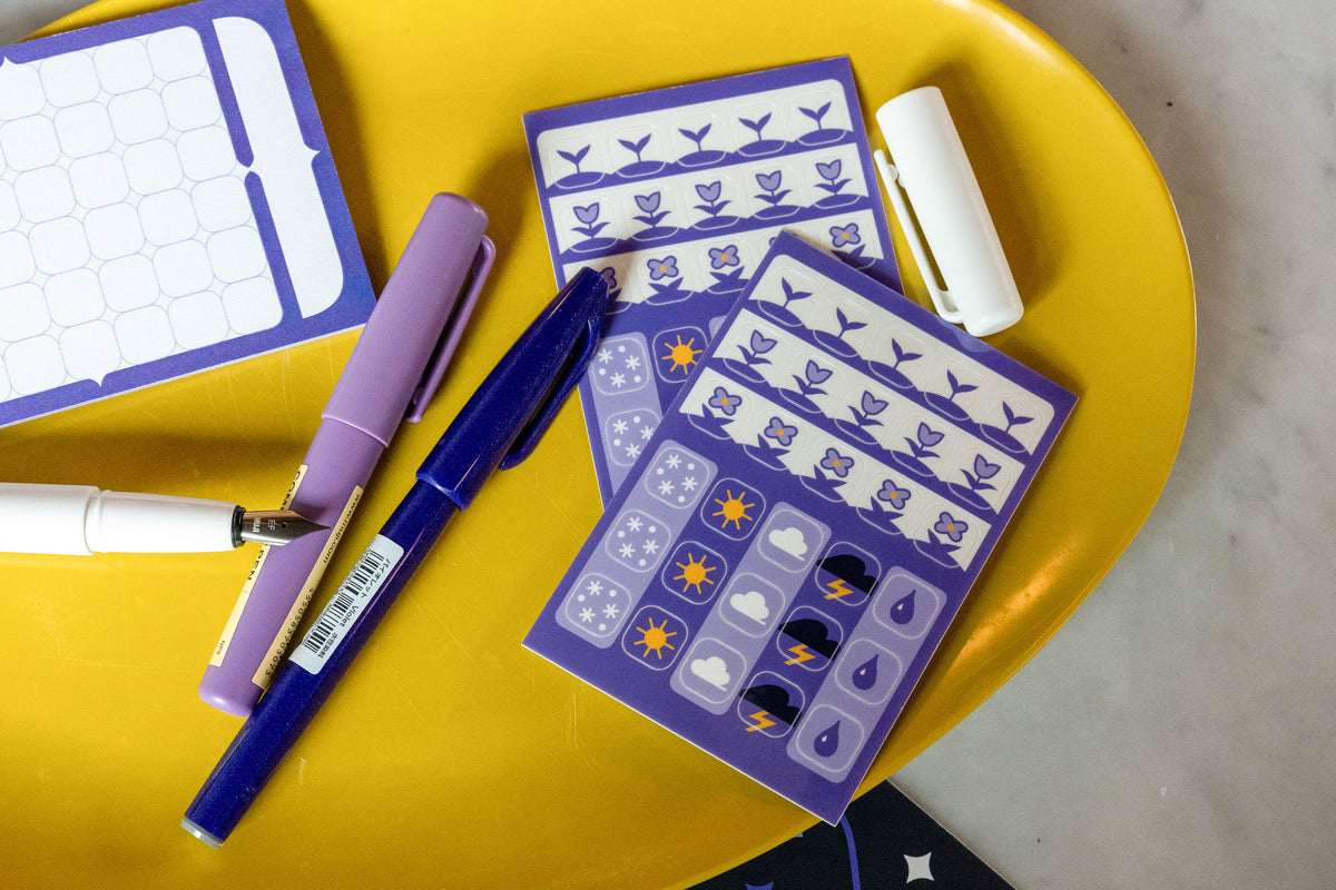 The Speedwell Icon Stickers photographed on a yellow tray with purple pens against a white marble background. The Calendar Sticky Note peeks in from left of frame.