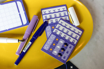The Speedwell Icon Stickers photographed on a yellow tray with purple pens against a white marble background. The Calendar Sticky Note peeks in from left of frame.