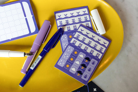 The Speedwell Icon Stickers photographed on a yellow tray with purple pens against a white marble background. The Calendar Sticky Note peeks in from left of frame.