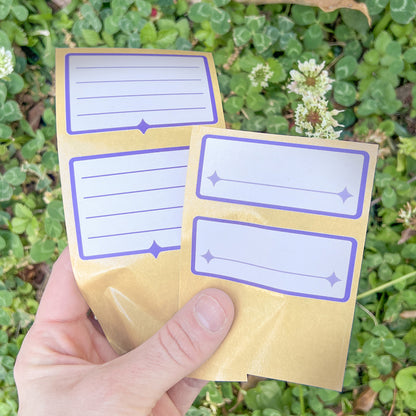 A hand holds an array of the white and indigo Nameplate Stickers against green clover and white clover blossoms.
