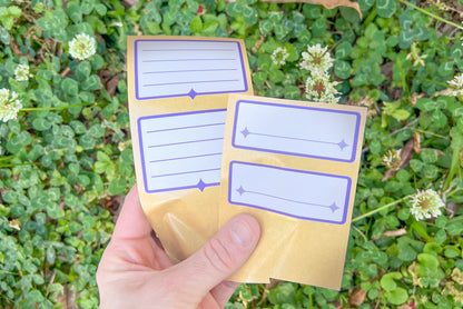 A hand holds an array of the white and indigo Nameplate Stickers against green clover and white clover blossoms.
