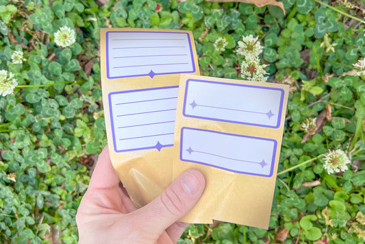 A hand holds an array of the white and indigo Nameplate Stickers against green clover and white clover blossoms.