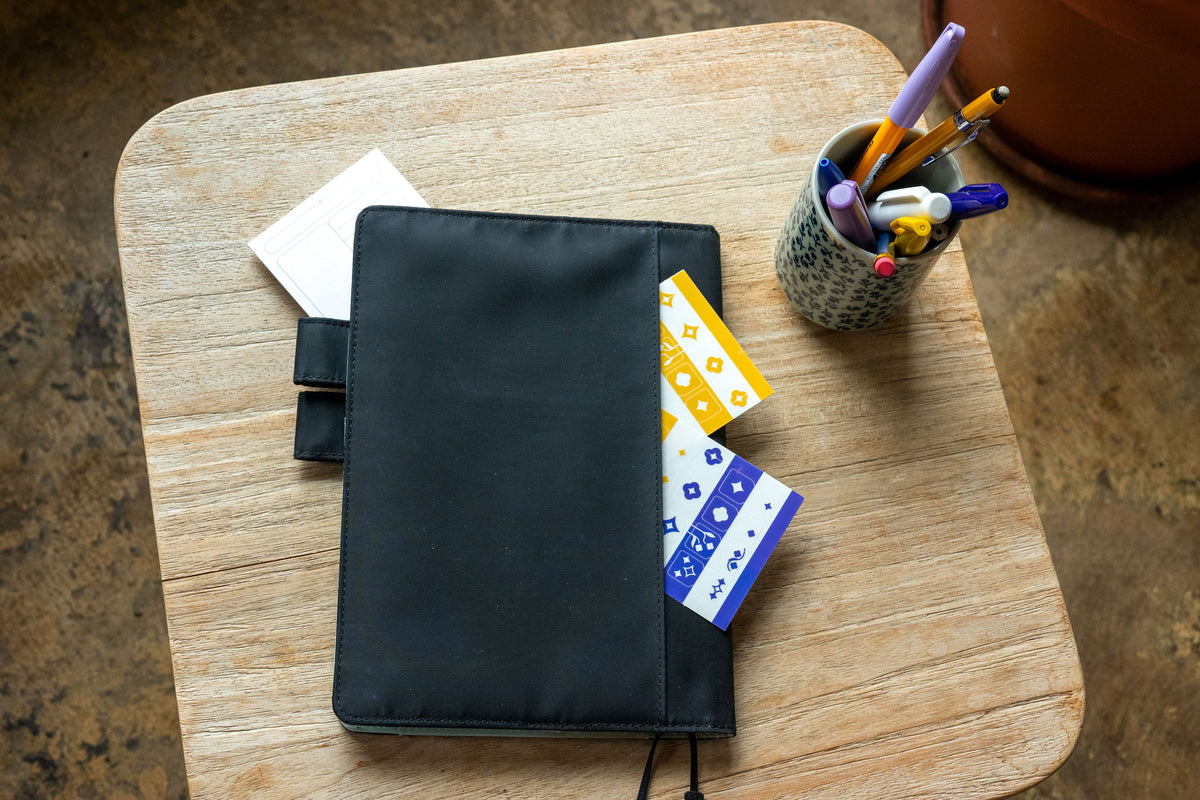 A planner with a black fabric cover lays on a wooden side table. The Archivist Sticky Note and Tab Divider Stickers are tucked into the the planner's back pocket. A pencil cup full of purple, yellow, and blue pencils sits to the right.