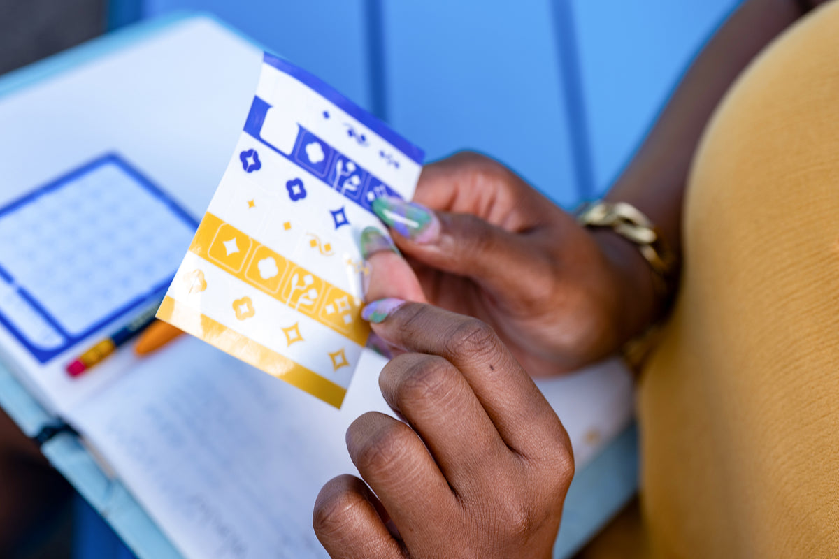 A close-up of a person's hands peeling a Tab Divider Sticker off its backer to place inside their notebook. The calendar sticky note can be seen inside the notebook.