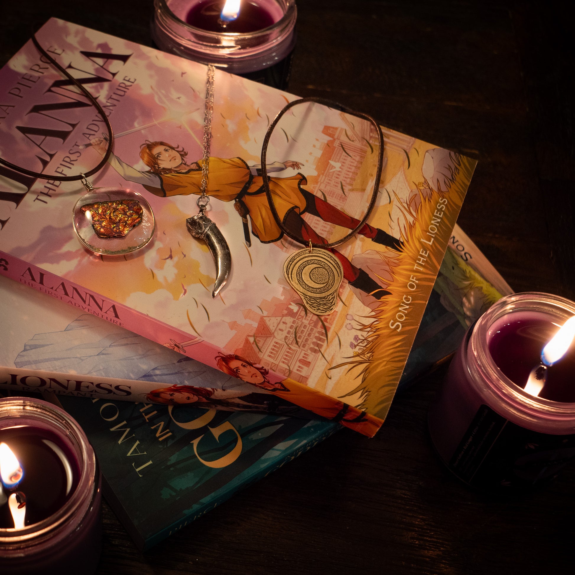 three necklaces - the badger claw, the ember glass, and the pregnancy charm - are laid on top of a pile of Alanna books, with candles surrounding.