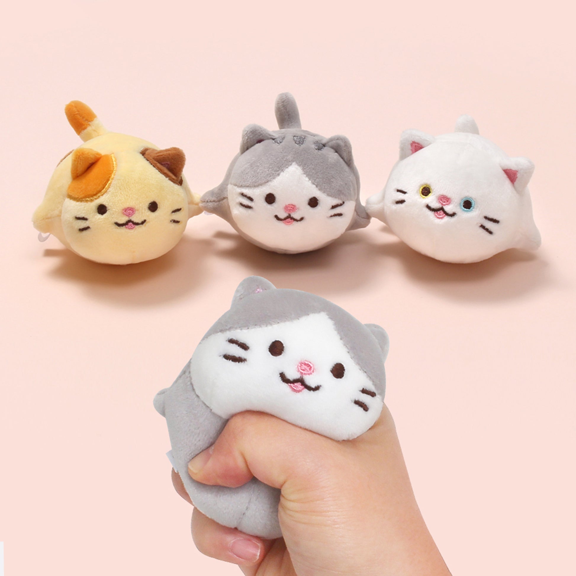 The whole cat trio: Mackerel, Odd, and Cheese, together on pastel pink background. In the foreground a hand squishes Mackerel. 