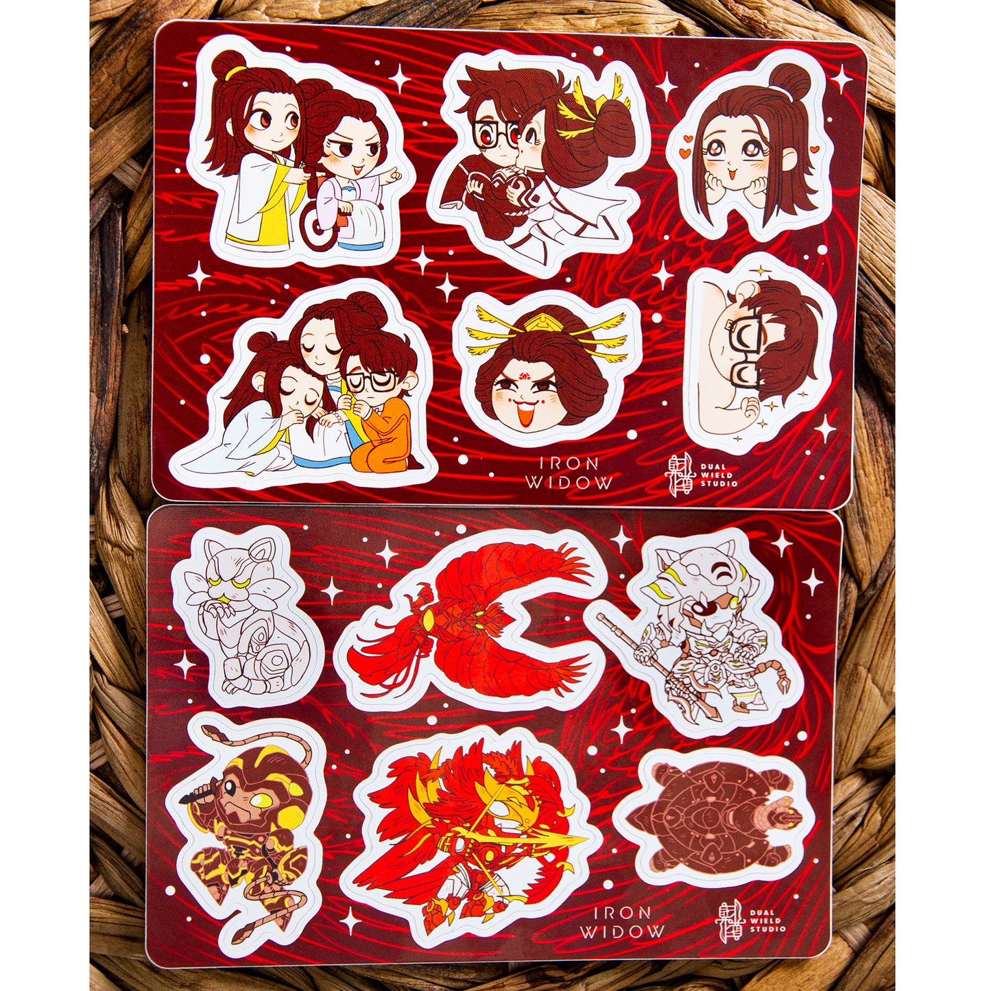 Iron Widow: Trio Sticker Sheet and Iron Widow: Mecha Sticker Sheet resting together on a warm brown wicker background.