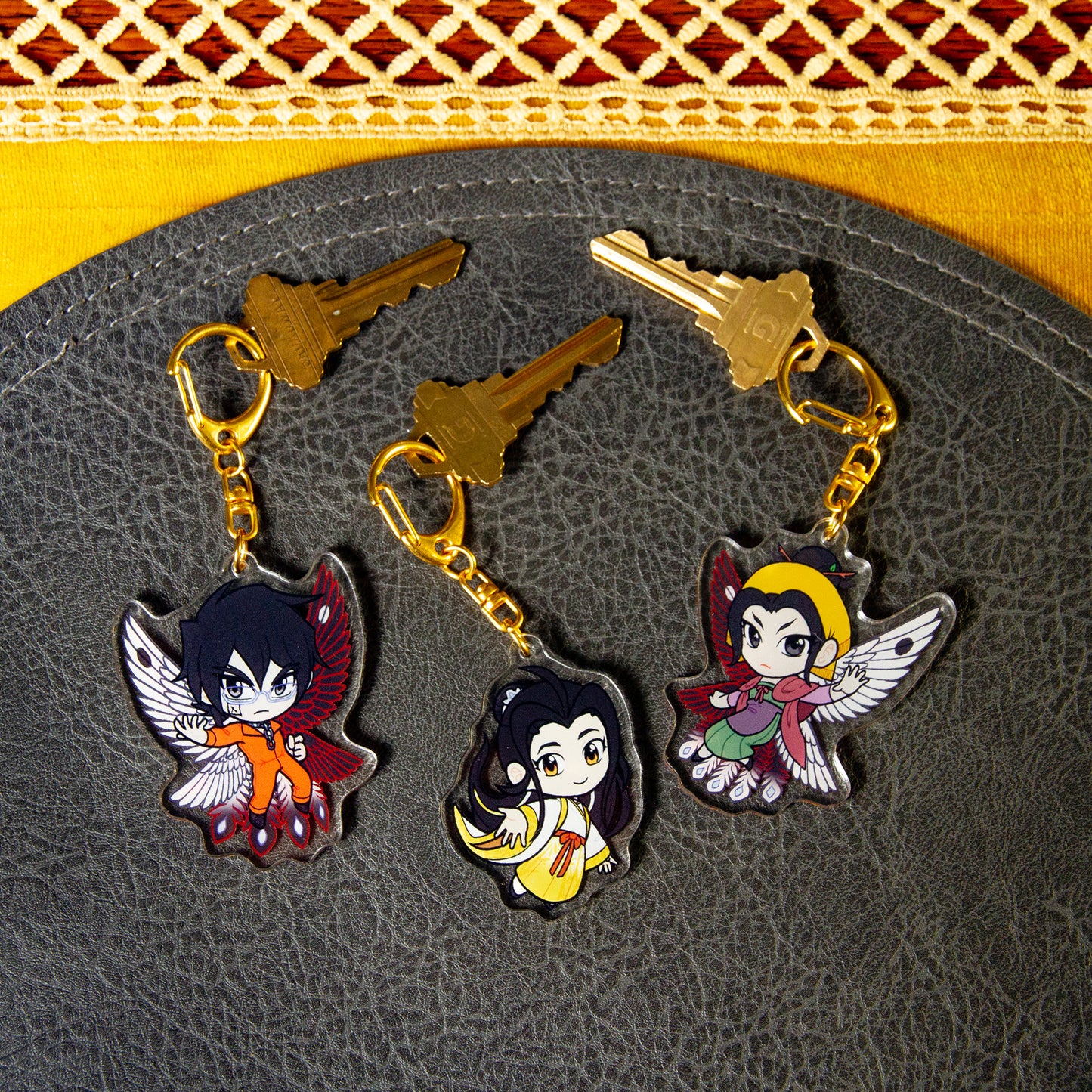 All three Iron Widow character charms side two, attached to house keys on a black pebbled leather mat.