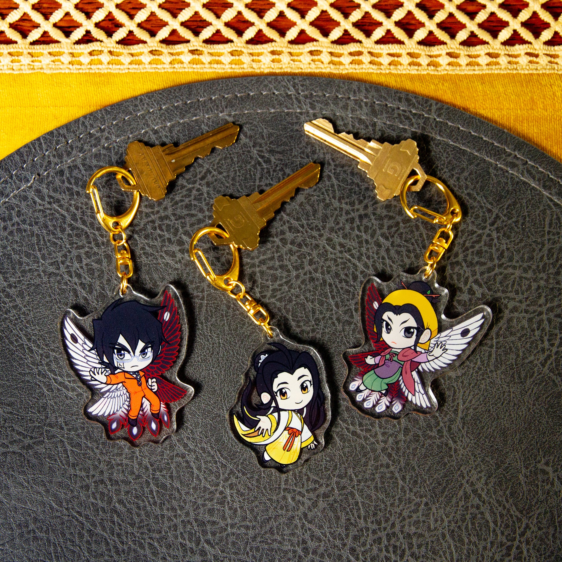 All three Iron Widow character charms side two, attached to house keys on a black pebbled leather mat.