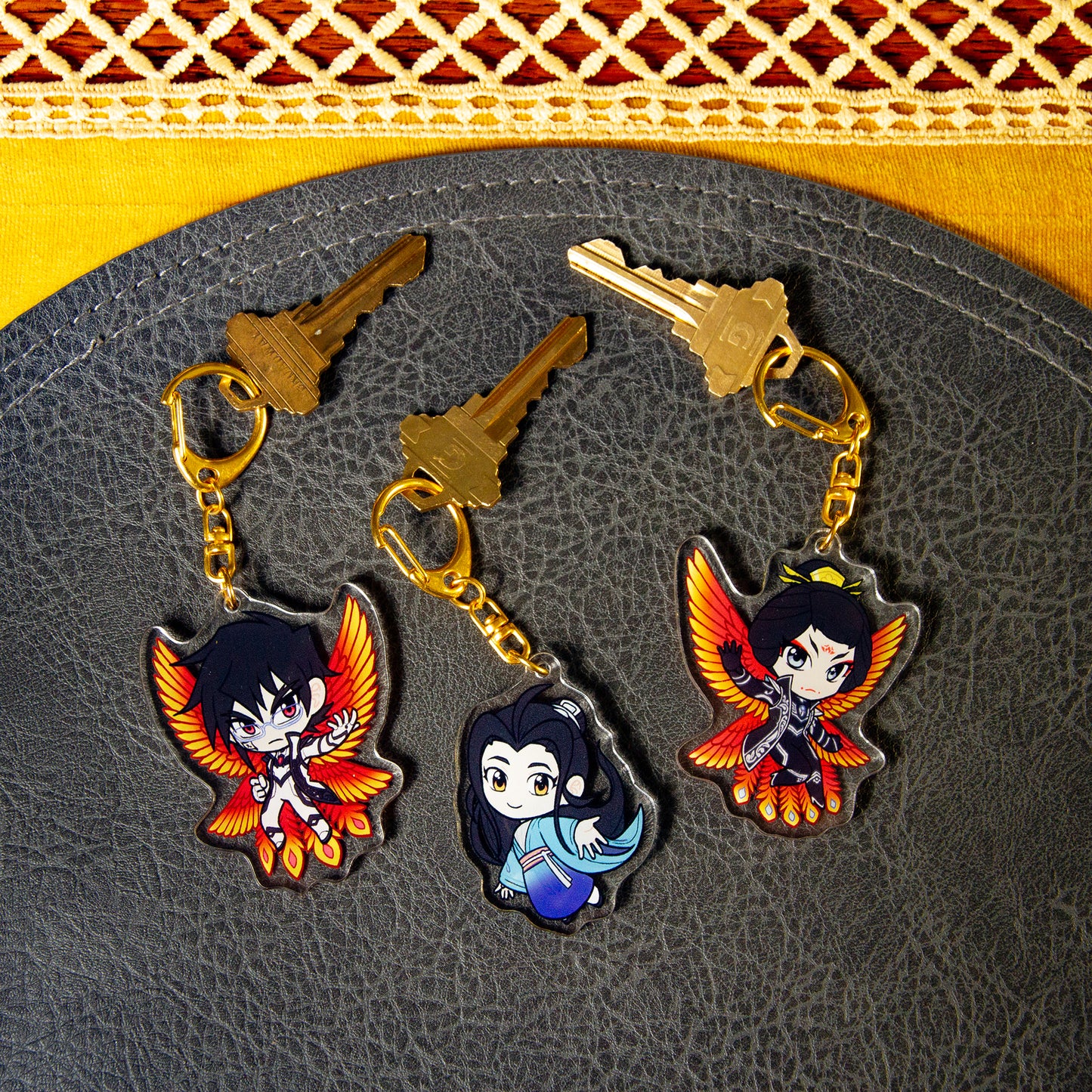 All three Iron Widow character charms side one, attached to house keys on a black pebbled leather mat.