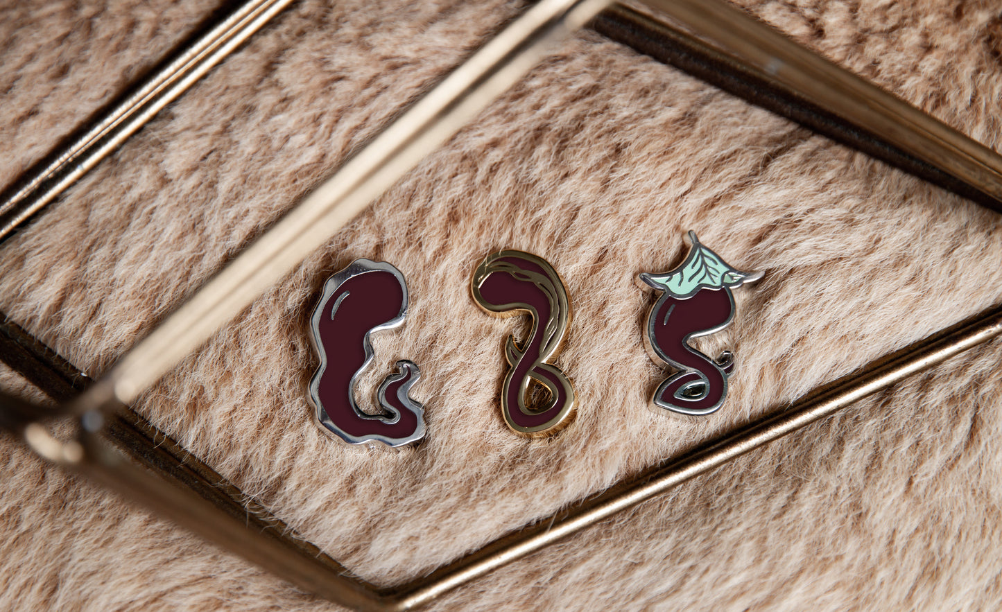 All Darking enamel pins (Jelly, Gold-Streak, and Leaf) on light brown fur surrounded by gold geometric decorative object.