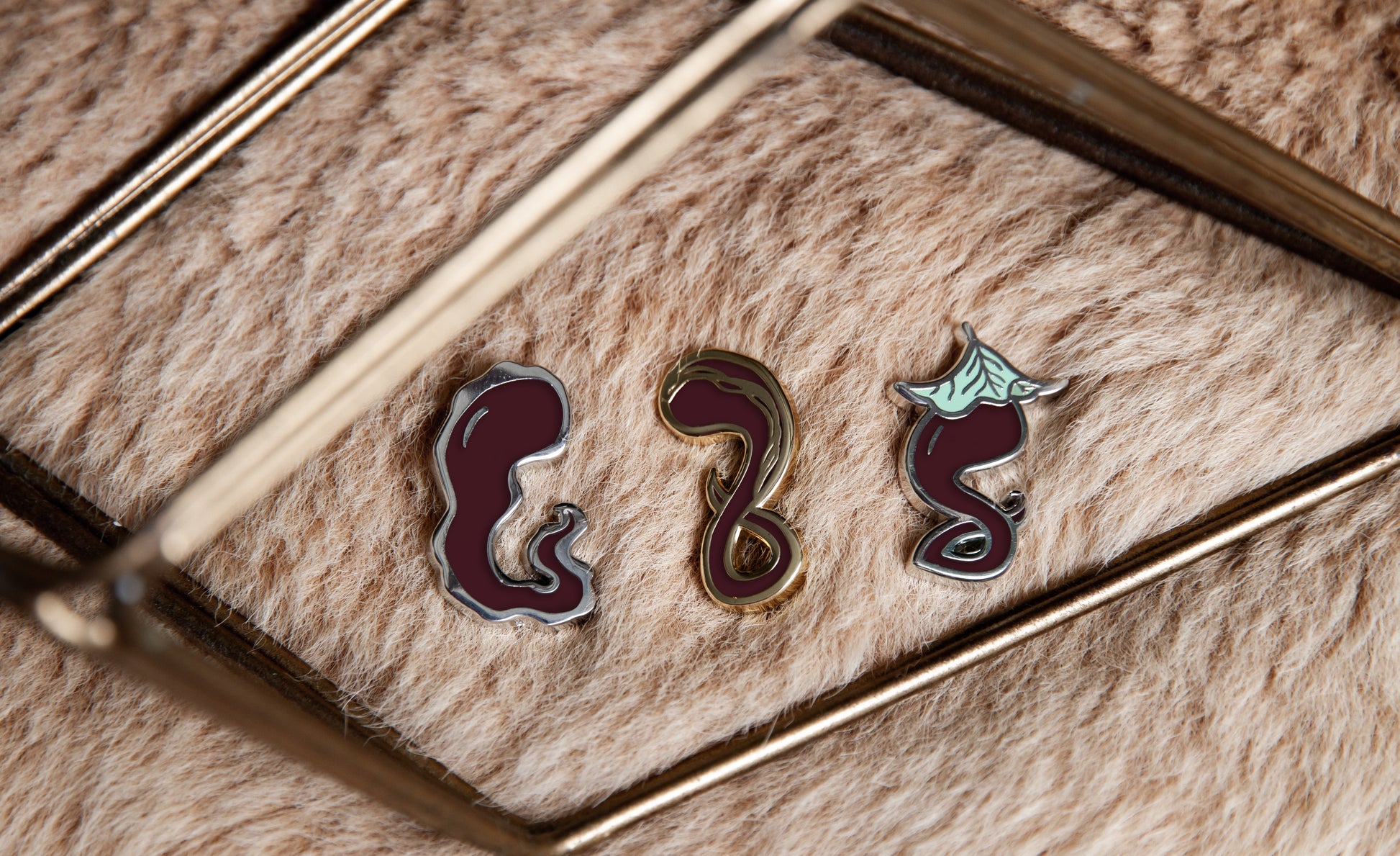 All Darking enamel pins (Jelly, Gold-Streak, and Leaf) on light brown fur surrounded by gold geometric decorative object.
